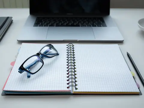 An open notebook and glasses in front of a laptop