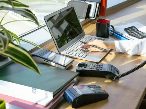 A sunny desk by a window with a laptop, a desk phone and a notebook